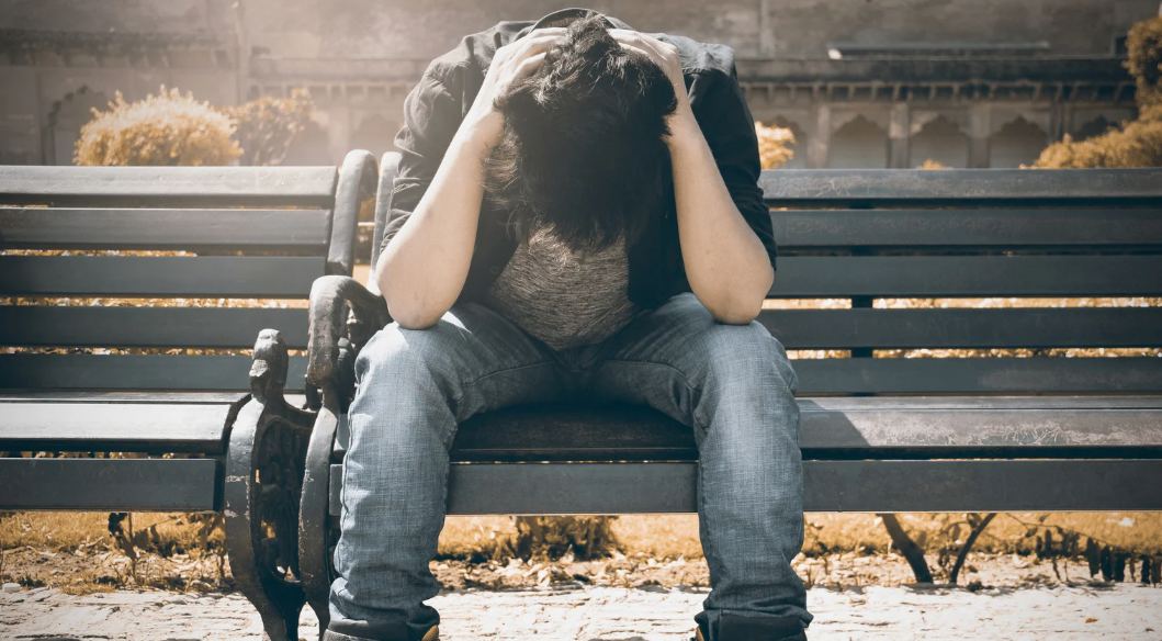 Ein junger Mann sitzt erschöpft mit dem Kopf in den Händen auf einer Parkbank